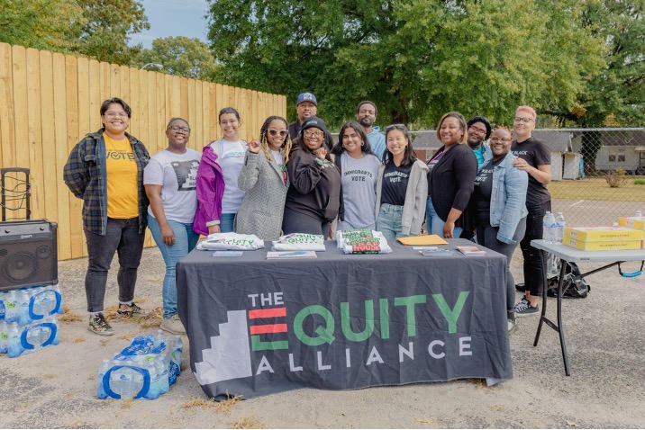 A group of people standing behind a table with a tablecloth that reads, "The Equity Alliance." The people are multiracial and smiling with their arms around each other. 
