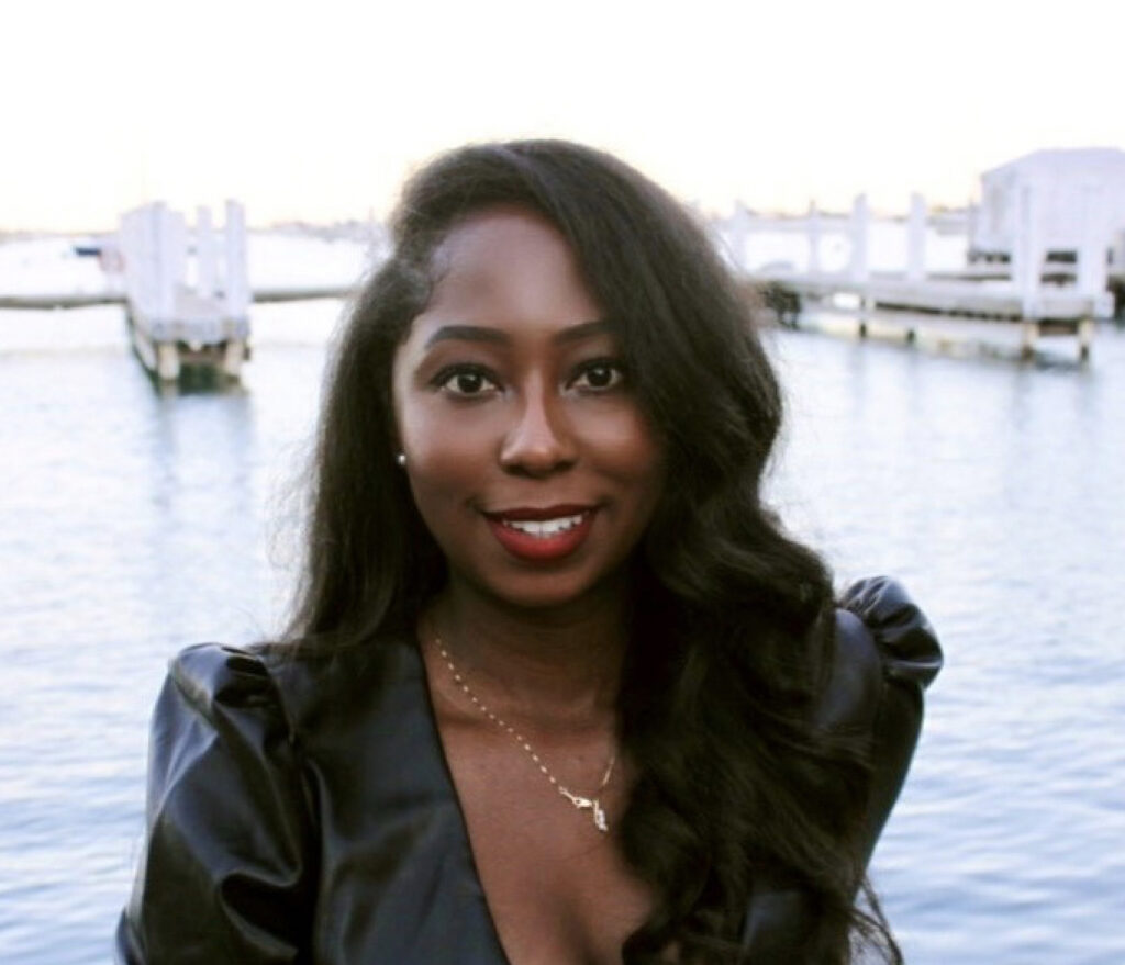 A Black woman wearing a Black shirt, smiling, standing in front of a body of water